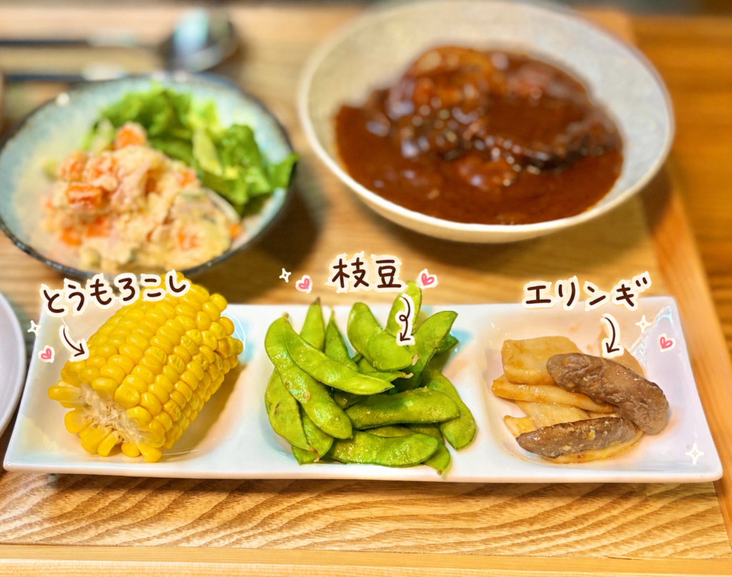 とうもろこし、枝豆、エリンギのオイスターソース炒めを横長の白い皿に盛りつけ、手書き風の可愛い文字で3品を紹介した副菜プレートの写真