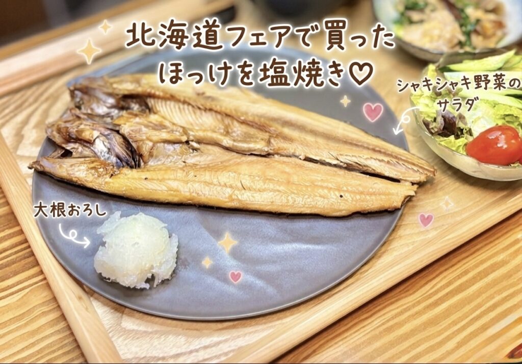 北海道フェアで買った大きなほっけを塩焼きにした、旦那キッチンの和食晩ごはん写真です。グレーのお皿に香ばしく焼かれたほっけの開きがどーんと盛りつけられ、横にはさっぱり食べられる大根おろしが添えられています。木製トレーの上には、シャキシャキ野菜のサラダやニラと豚バラ椎茸の出汁卵とじも並び、焼き魚を中心にした家庭的な和食献立の雰囲気が伝わります。写真には「北海道フェアで買ったほっけを塩焼き」「大根おろし」「シャキシャキ野菜のサラダ」という可愛い手書き風の文字入れがあり、ほっけの塩焼き献立、焼き魚の晩ごはん、和食のおうちごはん、旦那キッチンの記事