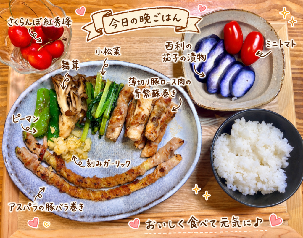 アスパラの豚バラ巻き、薄切り豚ロース肉の青紫蘇巻き、小松菜、ピーマン、舞茸、刻みガーリック、西利の茄子の漬物、ミニトマト、さくらんぼ紅秀峰、白ごはんを木のトレーに並べた晩ごはん献立写真。手書き風の文字で料理名を可愛く紹介している。